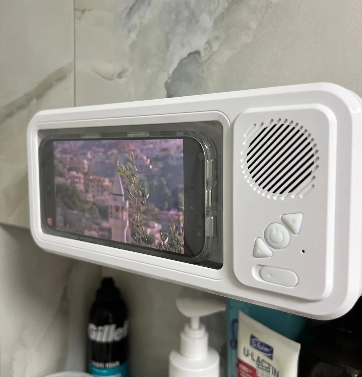 Actual white phone holder mounted on a bathroom wall, with phone inside and Bluetooth speaker at right